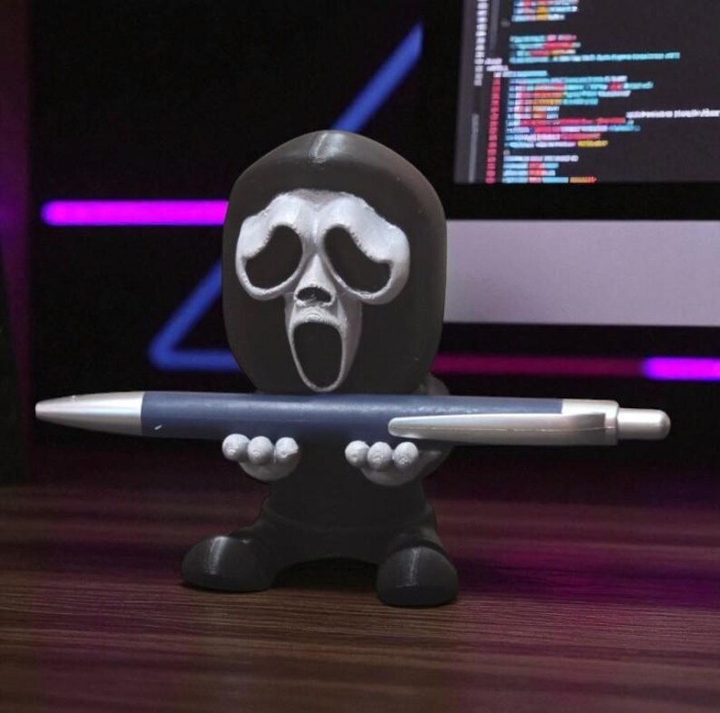 Puede incluir: Un portal&aacute;pices negro con forma de m&aacute;scara de Ghostface de las pel&iacute;culas Scream, sosteniendo un bol&iacute;grafo azul. El portal&aacute;pices tiene una cara blanca y est&aacute; sobre una superficie de madera.