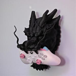 May include: A black dragon head wall mount holding a white and pink Xbox controller. The dragon's head is detailed with textured scales and sharp features. The controller has pink thumbsticks and blue accents.