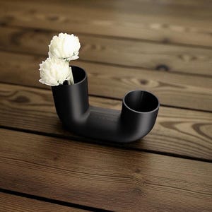 May include: A black, modern vase with a unique, curved design. Two white flowers are placed in one of the vase's openings. The vase is made of a matte material and sits on a wooden surface.