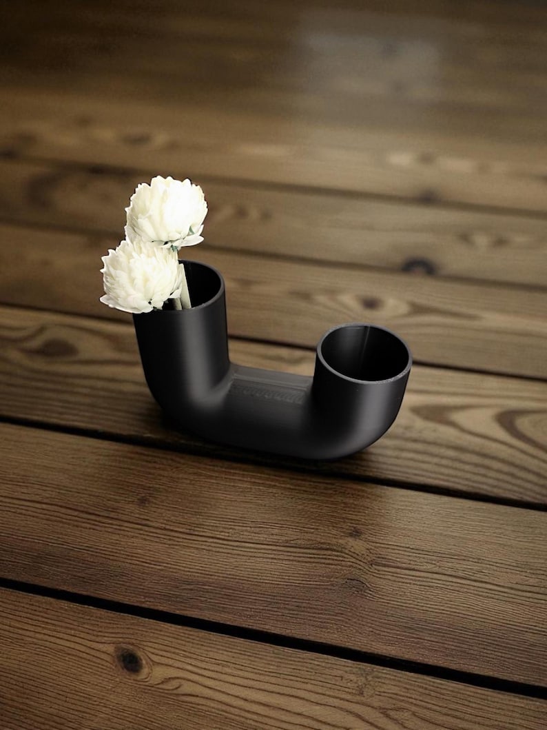May include: A black, modern vase with a unique, curved design. Two white flowers are placed in one of the vase's openings. The vase is made of a matte material and sits on a wooden surface.