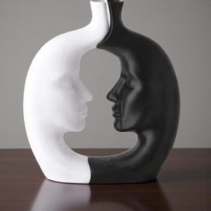 K&ouml;nnte beinhalten: Eine dekorative Vase in einem einzigartigen Design, das zwei einander zugewandte menschliche Gesichter zeigt. Eine Seite ist wei&szlig;, die andere ist schwarz. Die Vase hat ein gebogenes, offenes Design, wobei sich die Gesichter in der Mitte treffen. Die Vase ist aus Keramik.