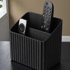 Op de afbeelding: Een zwarte, getextureerde afstandsbediening organizer met verticale geribbelde details. Twee afstandsbedieningen zijn zichtbaar, een zilverkleurige en een zwarte. De organizer is verdeeld in twee compartimenten en staat op een donkere ondergrond.
