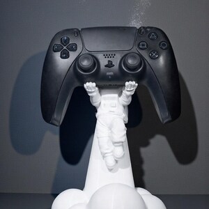 Op de afbeelding: Een zwarte PlayStation 5-controller die wordt vastgehouden door een wit astronautenfiguur. Het figuur staat op een wolkvormige basis. De controller heeft meerdere knoppen en richtingspads. De achtergrond is effen grijs.
