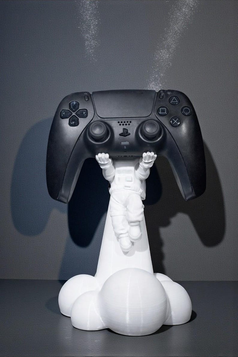 Op de afbeelding: Een zwarte PlayStation 5-controller die wordt vastgehouden door een wit astronautenfiguur. Het figuur staat op een wolkvormige basis. De controller heeft meerdere knoppen en richtingspads. De achtergrond is effen grijs.