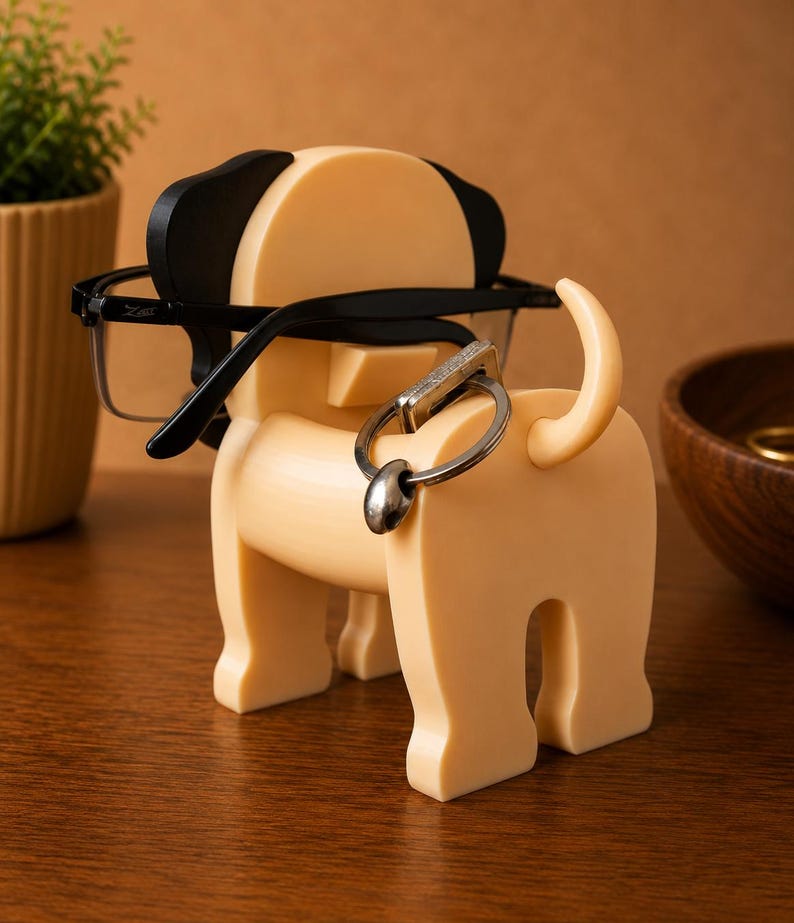 Pu&ograve; includere: Un portacavi a forma di cane, di colore beige chiaro, con dettagli neri sulle orecchie. Il supporto ha un portachiavi attaccato alla coda e tiene un paio di occhiali con montatura nera. Un oggetto decorativo per la casa o l'ufficio.