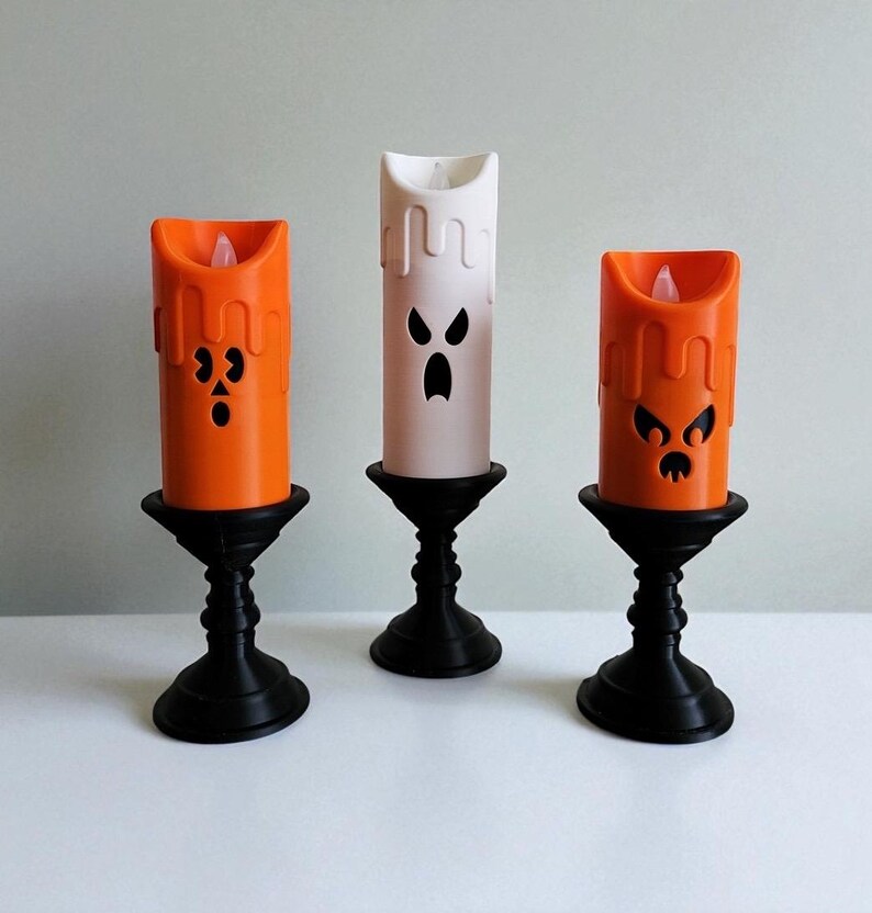 May include: Three Halloween-themed candle holders. Two orange candles and one white candle, each with a carved face. The candles sit on black, ornate stands. The orange candles have a dripping wax effect. The candles are lit with a soft glow.