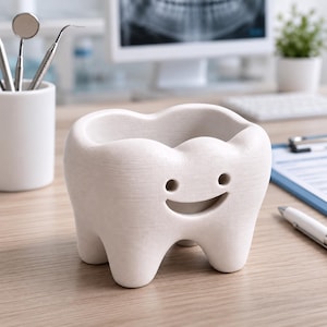 Może przedstawiać: Biały, ceramiczny pojemnik w kształcie zęba z uśmiechniętą twarzą. Pojemnik ma wydrążony wierzch i stoi na drewnianym biurku. W tle narzędzia dentystyczne i monitor komputera.