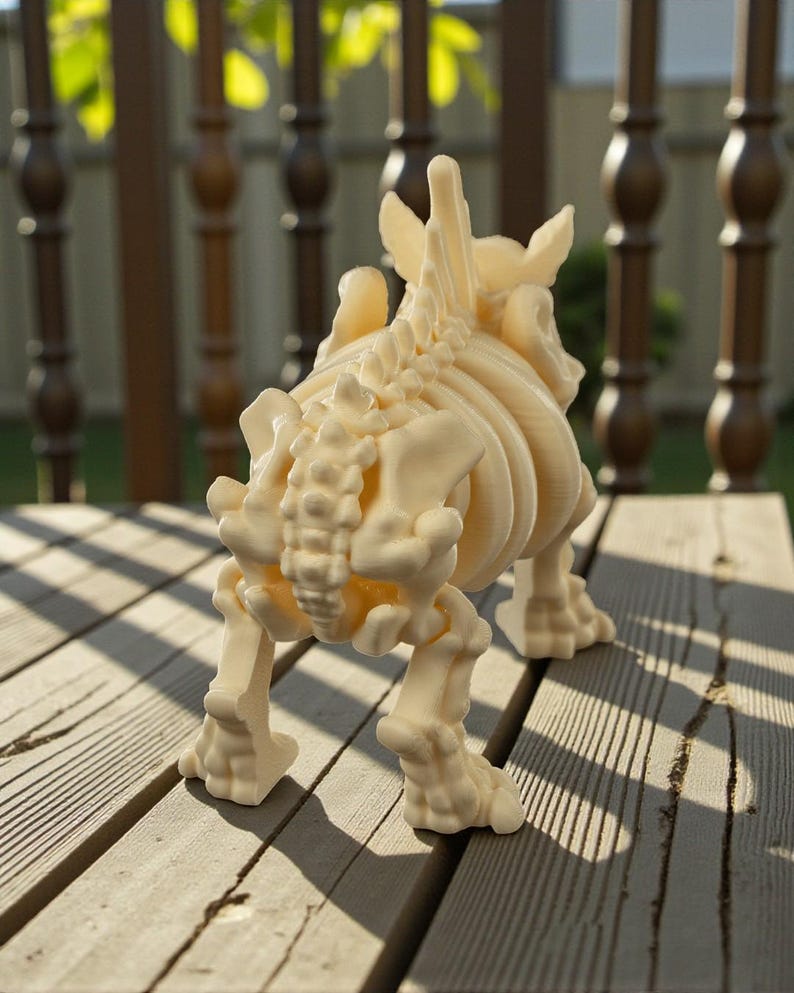 May include: A cream-colored, 3D-printed dinosaur skeleton figurine. The model is posed in a walking stance, showcasing the intricate details of the rib cage, spine, and leg bones. The figurine is displayed on a wooden surface.