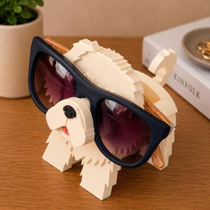 May include: A whimsical sunglasses holder shaped like a dog, crafted from off-white material. The dog has a black nose and a red tongue, holding a pair of navy blue sunglasses with purple lenses. The sunglasses have wooden arms.