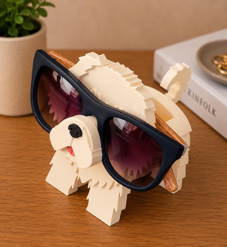 May include: A whimsical sunglasses holder shaped like a dog, crafted from off-white material. The dog has a black nose and a red tongue, holding a pair of navy blue sunglasses with purple lenses. The sunglasses have wooden arms.