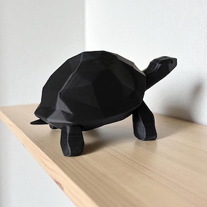 May include: A black geometric turtle figurine with a faceted design. The turtle is positioned on a light-colored wooden shelf against a white wall. The figurine has a modern, minimalist aesthetic.