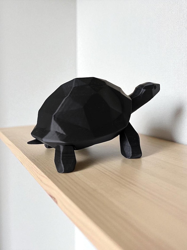 May include: A black geometric turtle figurine with a faceted design. The turtle is positioned on a light-colored wooden shelf against a white wall. The figurine has a modern, minimalist aesthetic.