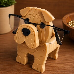 May include: A tan-colored dog-shaped eyeglasses holder with black-framed glasses. The holder is a detailed sculpture of a dog, designed to hold eyeglasses. The glasses are positioned as if the dog is wearing them.