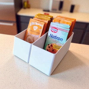 May include: Two white rectangular storage bins on a countertop. One bin contains packets labeled "Vanillinzucker", the other contains packets labeled "Dr. Oetker Natron". The packets are orange and blue.