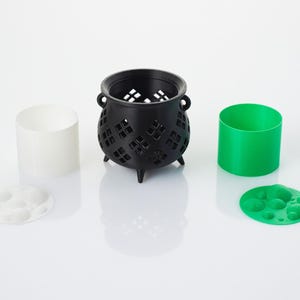 May include: A black cauldron with a decorative cut-out design, a white cylinder, and a green cylinder. The cauldron has three legs and is sitting on a white surface. There are also two small, round, white and green objects with raised circles on them.