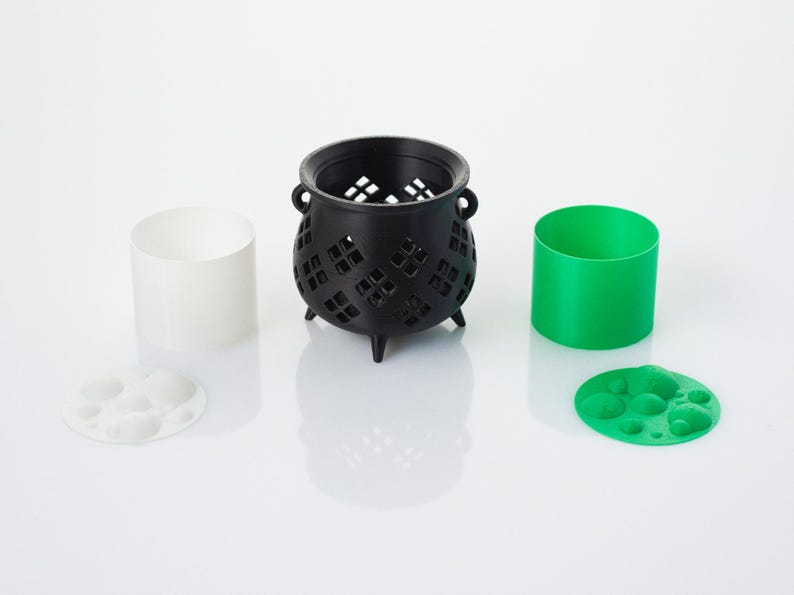 May include: A black cauldron with a decorative cut-out design, a white cylinder, and a green cylinder. The cauldron has three legs and is sitting on a white surface. There are also two small, round, white and green objects with raised circles on them.