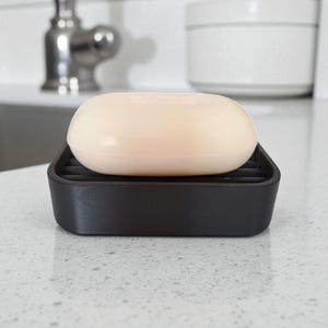 May include: A bar of light beige soap rests on a black soap dish with a slotted design. The soap dish is rectangular and sits on a light-colored countertop. The background includes a faucet and a white container.