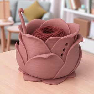 May include: A dusty rose-colored yarn bowl shaped like a rose, with a ball of dark red yarn inside. A crochet hook is placed in the bowl. The bowl has three small holes on the side.