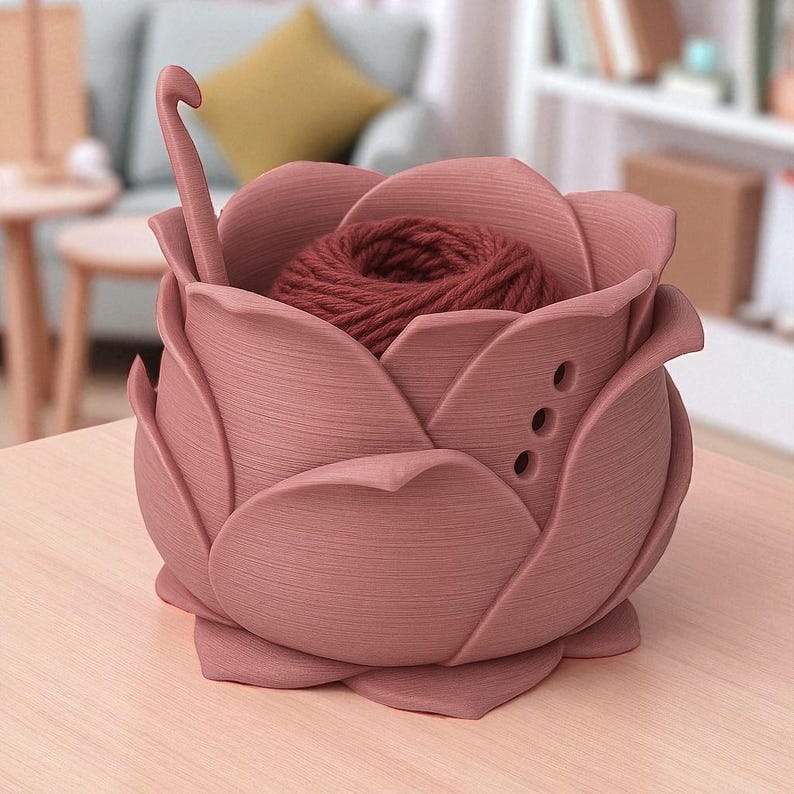 May include: A dusty rose-colored yarn bowl shaped like a rose, with a ball of dark red yarn inside. A crochet hook is placed in the bowl. The bowl has three small holes on the side.