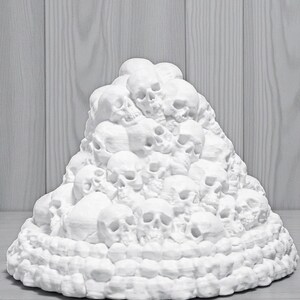 May include: A white sculpture of a mound of skulls. The skulls are stacked on top of each other, creating a textured, three-dimensional effect. The sculpture is set against a light gray wooden background.
