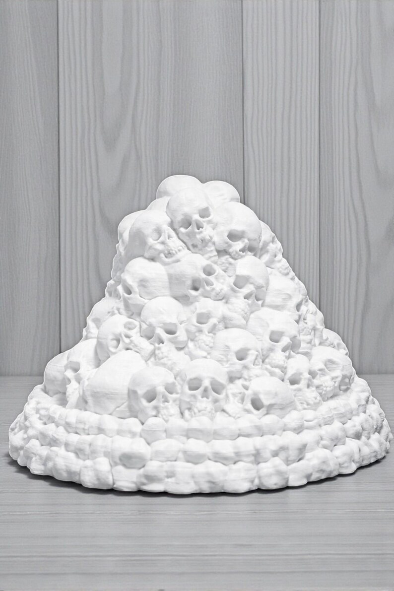 May include: A white sculpture of a mound of skulls. The skulls are stacked on top of each other, creating a textured, three-dimensional effect. The sculpture is set against a light gray wooden background.