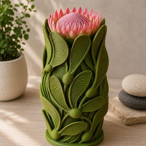 以下が含まれることがあります： スタイリッシュな緑色の花瓶。葉と種子の鞘をモチーフにした、詳細な植物デザインが施されています。花瓶には大きなピンク色のプロテアの花が入っています。複雑なデザインと自然な色合いが特徴の装飾品です。