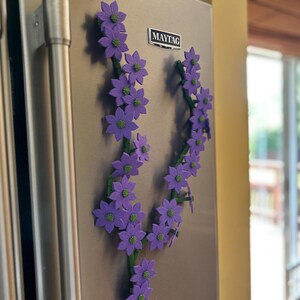 May include: Purple flower magnets arranged in a vine-like pattern, attached to a stainless steel refrigerator. The flowers have green centers and are set against a neutral background. A Maytag logo is visible.