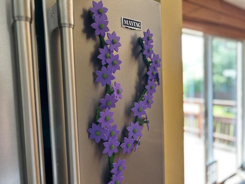 May include: Purple flower magnets arranged in a vine-like pattern, attached to a stainless steel refrigerator. The flowers have green centers and are set against a neutral background. A Maytag logo is visible.