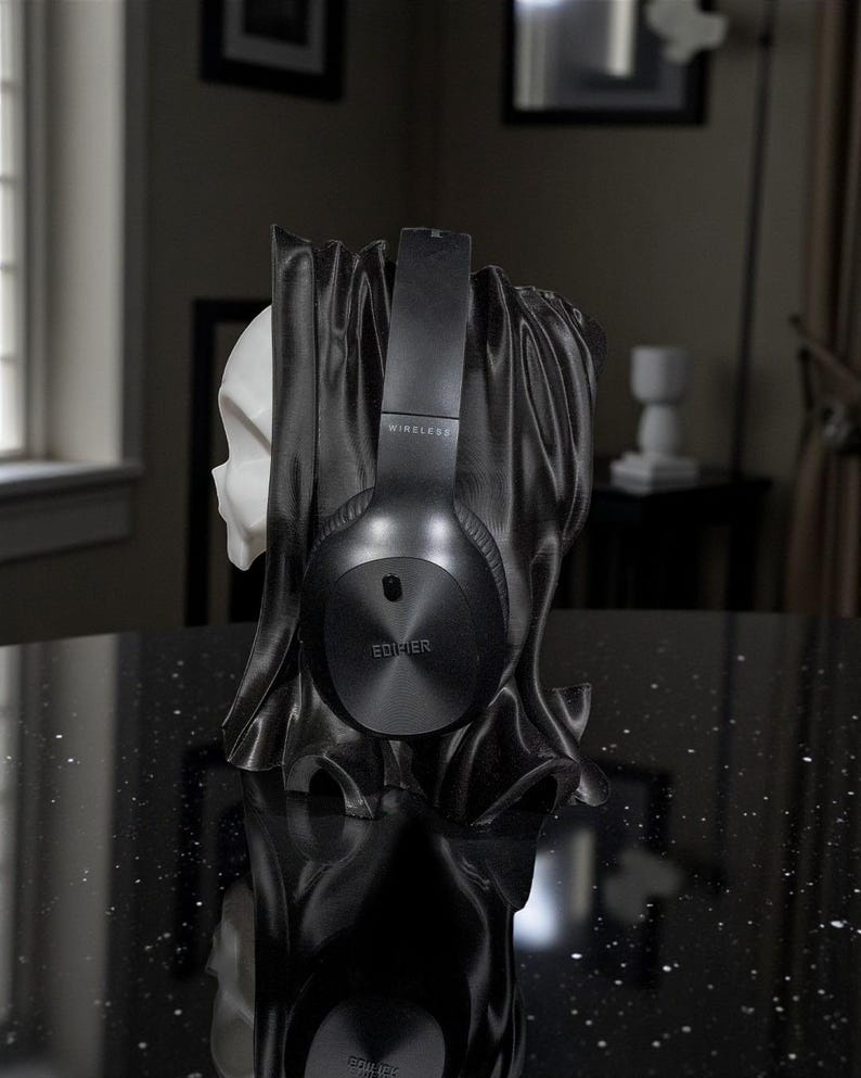 May include: Black wireless Edifier headphones resting on a black skull-shaped headphone stand. The stand has a draped fabric design. The headphones are centered, with the word "WIRELESS" visible on the headband.