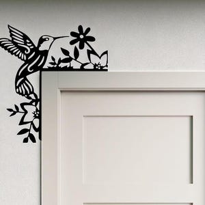 May include: Black metal hummingbird and floral corner decoration. The hummingbird is depicted in flight, with detailed wing and feather designs. The flowers and leaves are also cut from black metal, creating a decorative accent for a door frame or wall.