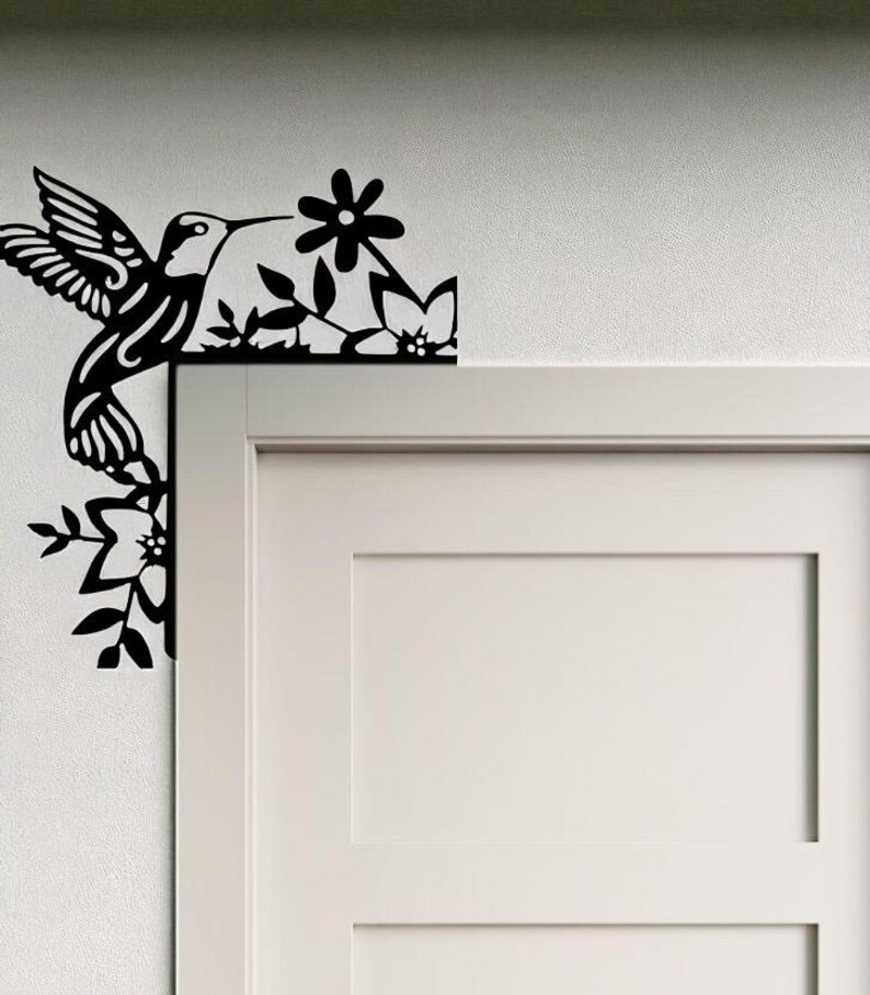 May include: Black metal hummingbird and floral corner decoration. The hummingbird is depicted in flight, with detailed wing and feather designs. The flowers and leaves are also cut from black metal, creating a decorative accent for a door frame or wall.