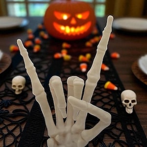 May include: A Halloween table setting featuring a carved pumpkin with a glowing face, a white skeleton hand making a rock-and-roll gesture, and scattered candy corn. Two small skull figurines are also present.
