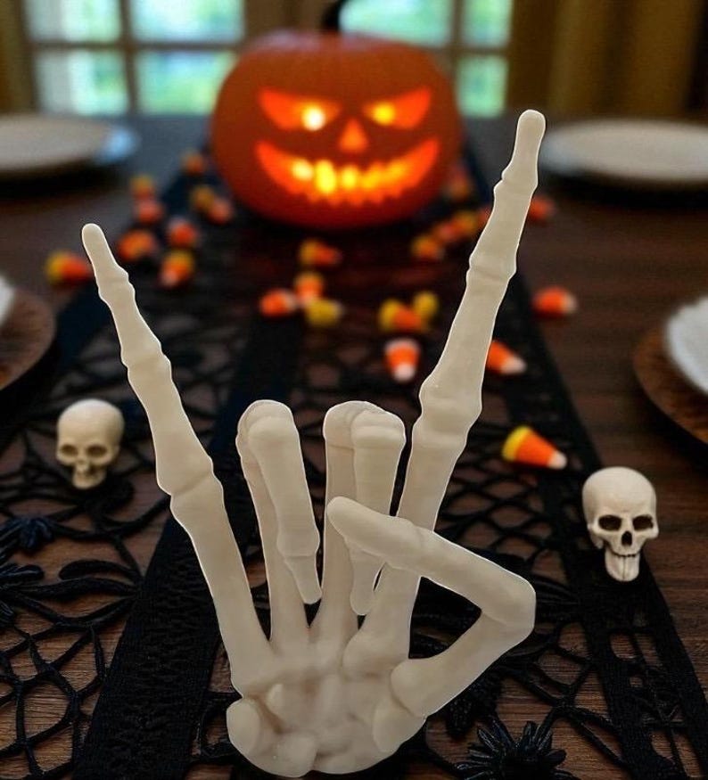 May include: A Halloween table setting featuring a carved pumpkin with a glowing face, a white skeleton hand making a rock-and-roll gesture, and scattered candy corn. Two small skull figurines are also present.