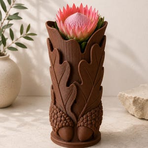 Op de afbeelding: Een bruine, decoratieve vaas met een eikenblad- en eikelontwerp, met een roze en rode protea-bloem. De vaas is gemaakt van een textuurrijk materiaal en heeft een gedetailleerde, naturalistische uitstraling. De bloem staat in volle bloei.