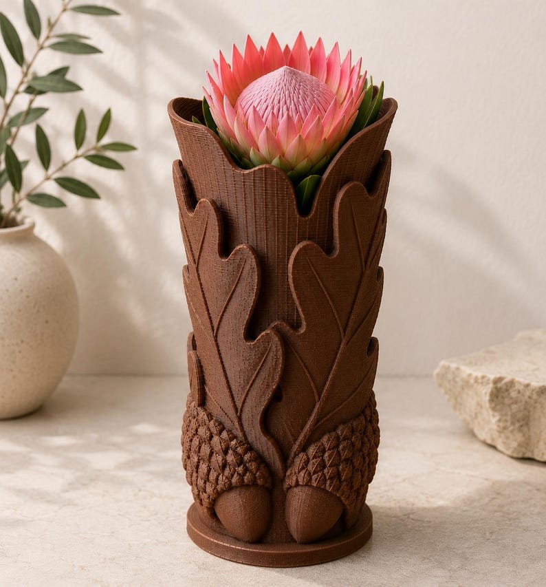Op de afbeelding: Een bruine, decoratieve vaas met een eikenblad- en eikelontwerp, met een roze en rode protea-bloem. De vaas is gemaakt van een textuurrijk materiaal en heeft een gedetailleerde, naturalistische uitstraling. De bloem staat in volle bloei.