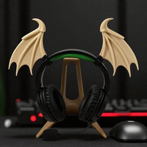 May include: A black gaming headset with a green accent sits on a beige stand, with beige bat wings on either side. A black mouse is to the right. The headset has a microphone and is designed for gaming.