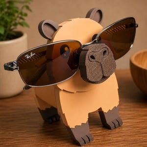 May include: A whimsical, tan capybara-shaped sunglasses holder with brown accents. The holder features a detailed nose and ears, and is displaying a pair of brown-tinted sunglasses with a silver frame. The sunglasses are resting on the capybara's nose.