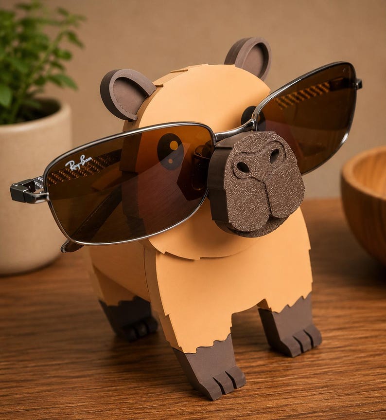 May include: A whimsical, tan capybara-shaped sunglasses holder with brown accents. The holder features a detailed nose and ears, and is displaying a pair of brown-tinted sunglasses with a silver frame. The sunglasses are resting on the capybara's nose.