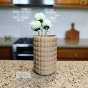 Pu&ograve; includere: Un vaso beige strutturato con un design arrotondato a bolle, contenente tre fiori bianchi con steli verdi. Il vaso cilindrico &egrave; posto su un piano di lavoro maculato, per l'arredamento della casa.