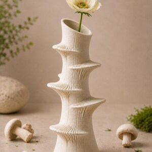 May include: A cream-colored, textured ceramic vase with a unique spiral design, holding a single white flower with a yellow center. The vase is surrounded by mushrooms and greenery, creating a natural, earthy aesthetic.