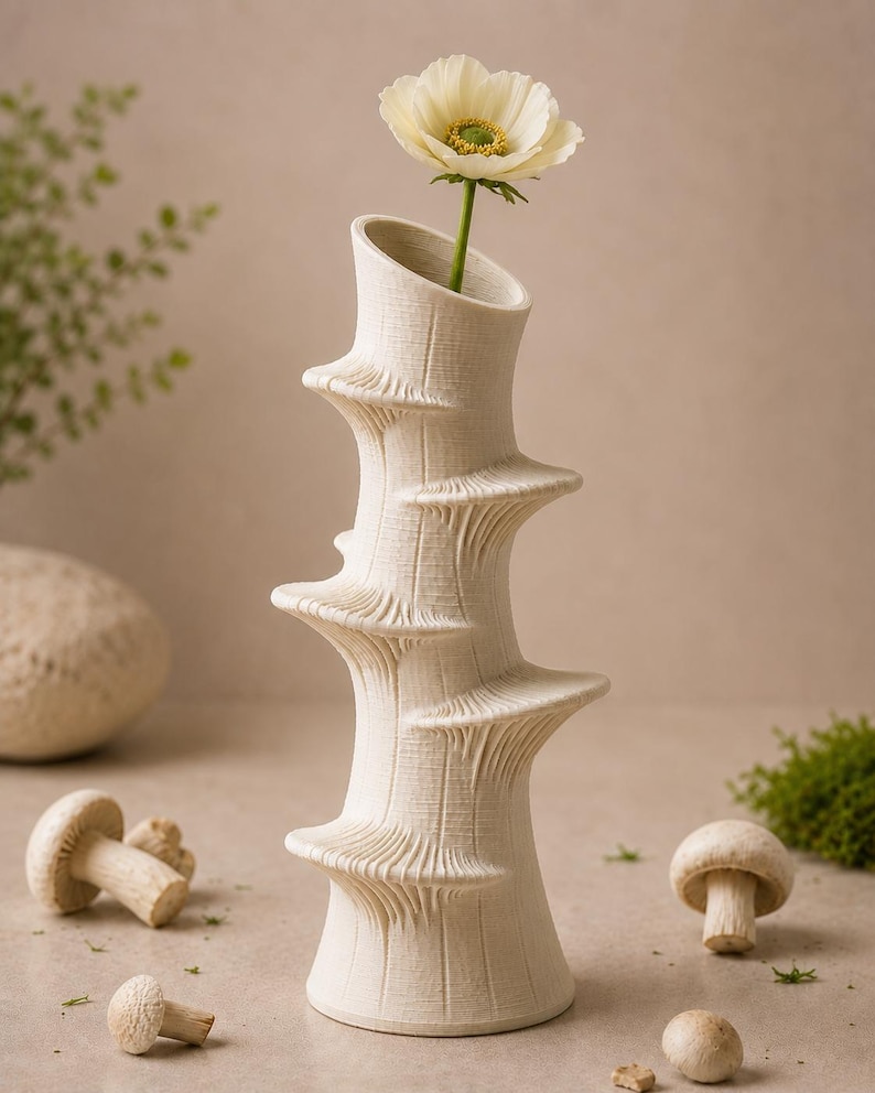 May include: A cream-colored, textured ceramic vase with a unique spiral design, holding a single white flower with a yellow center. The vase is surrounded by mushrooms and greenery, creating a natural, earthy aesthetic.