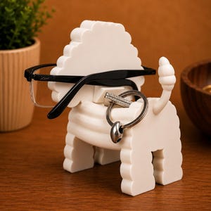 May include: A white, lion-shaped eyeglasses and key holder. The holder has a textured mane and tail, with a pair of black-framed glasses resting on top and a silver key ring hanging from the neck. The holder is on a wooden surface.