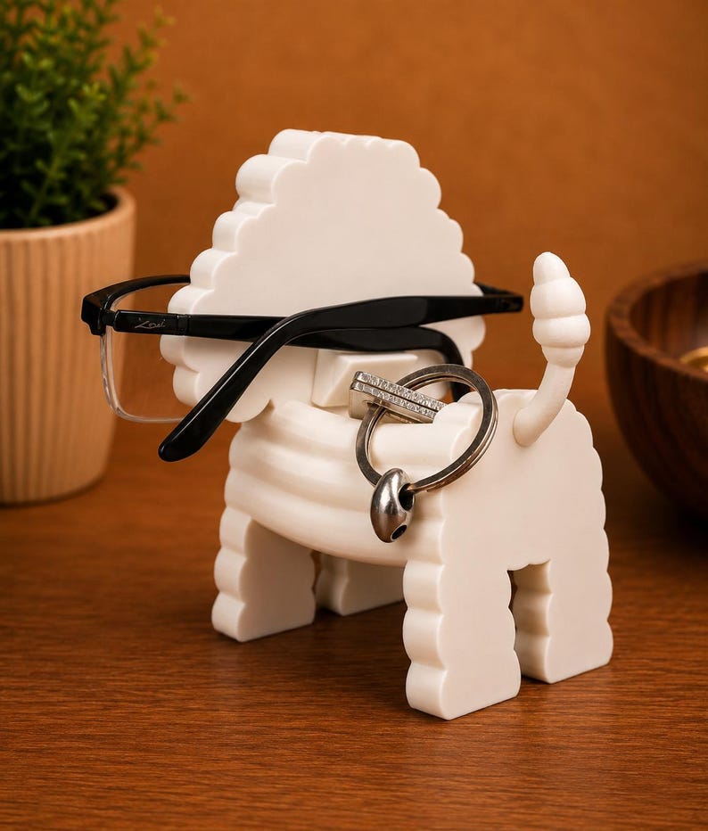 May include: A white, lion-shaped eyeglasses and key holder. The holder has a textured mane and tail, with a pair of black-framed glasses resting on top and a silver key ring hanging from the neck. The holder is on a wooden surface.