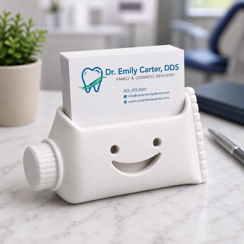 Pu&ograve; includere: Un portacarte bianco a forma di tubetto di dentifricio, con un design a faccina sorridente. Il supporto contiene una pila di biglietti da visita bianchi con il testo "Dr. Emily Carter, DDS" e informazioni di contatto.