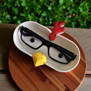 Op de afbeelding: Een witte keramische tray met een bril met zwart montuur, een rood decoratief stuk en een geel hartvormig object. De tray staat op een houten oppervlak, met een groene, bladerrijke achtergrond.