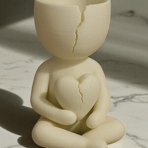 May include: A cream-colored figurine of a person seated with legs crossed, holding a cracked heart. The head of the figurine is also cracked. The sculpture is a minimalist design, possibly a decorative item or art piece.