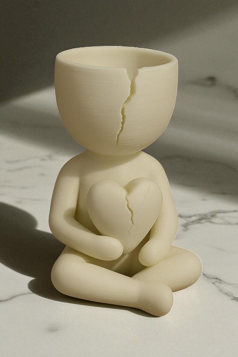May include: A cream-colored figurine of a person seated with legs crossed, holding a cracked heart. The head of the figurine is also cracked. The sculpture is a minimalist design, possibly a decorative item or art piece.