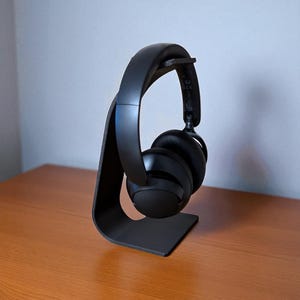 May include: A black headphone stand with a curved design, holding a pair of black over-ear headphones. The stand is made of a solid material and sits on a wooden surface. The headphones are positioned on the stand, ready for use.