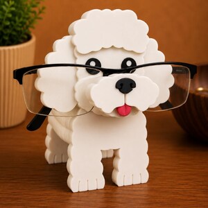 May include: A white, poodle-shaped eyeglass holder with black framed glasses. The holder has a black nose and eyes, and a pink tongue. The holder is made of a layered material and sits on a wooden surface.