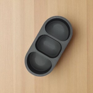 May include: A dark gray, three-sectioned serving tray with rounded compartments. The tray is made of a textured material and is positioned diagonally on a light wood surface. The compartments are oval-shaped.